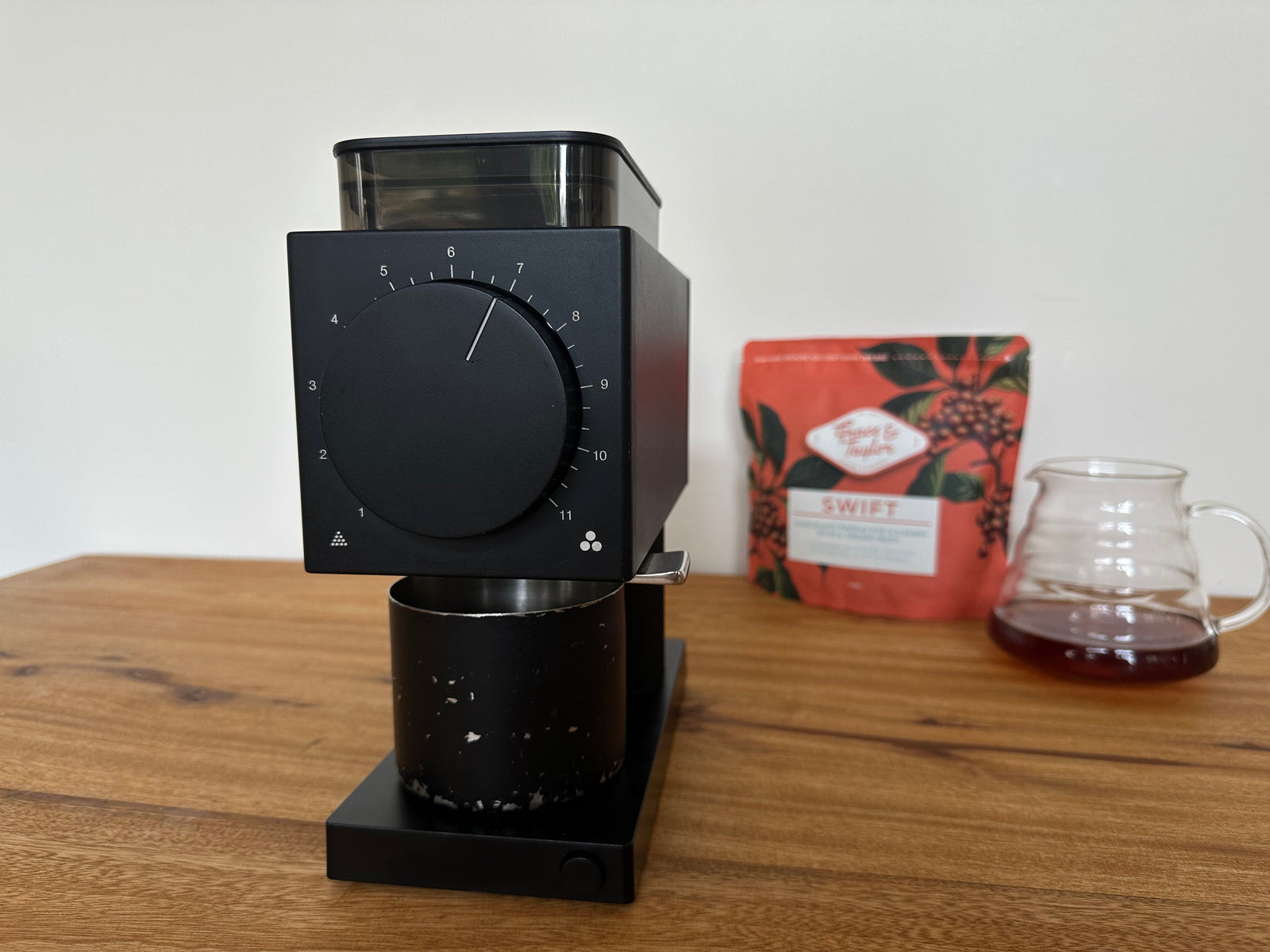 A coffee grinder sitting on a kitchen bench, with a bag of coffee and a pot of coffee in the background