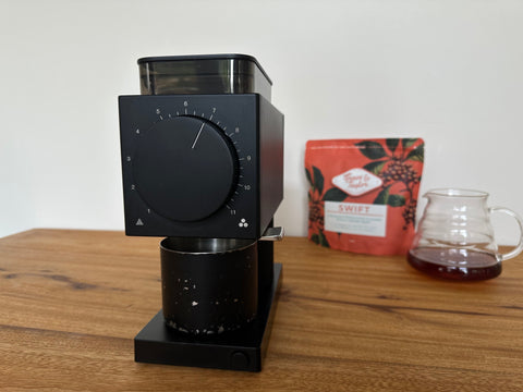A coffee grinder sitting on a kitchen bench, with a bag of coffee and a pot of coffee in the background